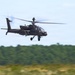 A military attack helicopter, consistent with an AH-64 Apache design, is captured in low-level flight against a light blue sky with a blurred tree line in the background. The motion blur on the rotor blades and background indicates the helicopter is moving at speed. The image appears to be taken with a panning technique, keeping the aircraft sharp while blurring the surroundings.