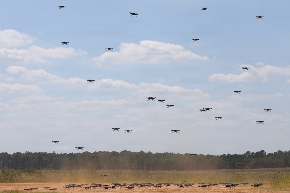 A large swarm of quadcopter drones is captured in various stages of flight over an open dirt field surrounded by a tree line. The drones range from ground level to high altitude, suggesting a mass launch or coordinated swarm exercise. The setting appears to be a military or research testing ground based on the terrain and scale of the operation.