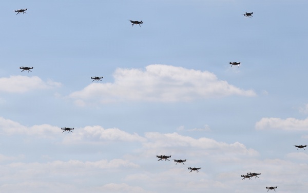 A large swarm of quadcopter drones is captured in various stages of flight over an open dirt field surrounded by a tree line. The drones range from ground level to high altitude, suggesting a mass launch or coordinated swarm exercise. The setting appears to be a military or research testing ground based on the terrain and scale of the operation.