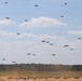 A large swarm of quadcopter drones is captured in various stages of flight over an open dirt field surrounded by a tree line. The drones range from ground level to high altitude, suggesting a mass launch or coordinated swarm exercise. The setting appears to be a military or research testing ground based on the terrain and scale of the operation.