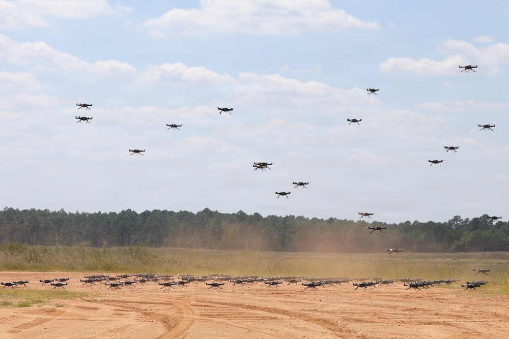 A large swarm of quadcopter drones is captured in various stages of takeoff and flight over a sandy open field. Dozens of drones are visible both on the ground and ascending into the sky, suggesting a coordinated mass launch exercise. The setting appears to be a military or testing facility surrounded by pine trees.