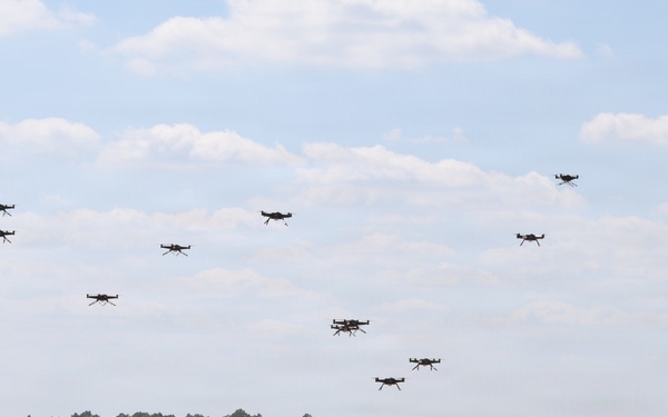 A large swarm of quadcopter drones is captured in various stages of takeoff and flight over a sandy open field. Dozens of drones are visible both on the ground and ascending into the sky, suggesting a coordinated mass launch exercise. The setting appears to be a military or testing facility surrounded by pine trees.