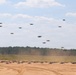 A large swarm of quadcopter drones is captured in various stages of takeoff and flight over a sandy open field. Dozens of drones are visible both on the ground and ascending into the sky, suggesting a coordinated mass launch exercise. The setting appears to be a military or testing facility surrounded by pine trees.