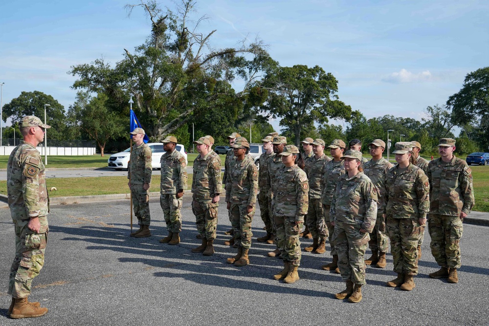 A military formation is taking place outdoors on a paved surface, with a senior officer or NCO addressing a group of approximately 20 uniformed personnel standing at attention. All individuals are wearing OCP (Operational Camouflage Pattern) uniforms with tan combat boots, consistent with U.S. Air Force or Space Force dress standards. A blue guidon flag is visible being carried by one of the formation members.