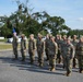 A military formation is taking place outdoors on a paved surface, with a senior officer or NCO addressing a group of approximately 20 uniformed personnel standing at attention. All individuals are wearing OCP (Operational Camouflage Pattern) uniforms with tan combat boots, consistent with U.S. Air Force or Space Force dress standards. A blue guidon flag is visible being carried by one of the formation members.