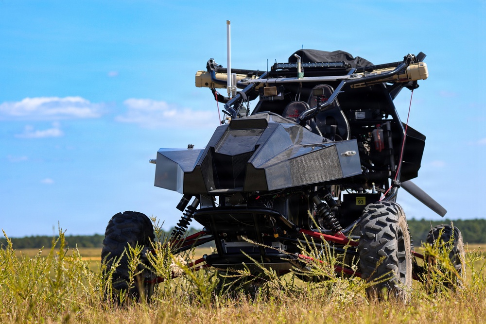 A heavily modified off-road utility vehicle (UTV/buggy) is stationary in an open grassy field under a clear blue sky. The vehicle has been extensively outfitted with what appears to be military or tactical equipment, including mounted systems on top, antennas, armor plating, and various electronic components. The vehicle appears to be an unmanned ground vehicle (UGV) or a remotely operated tactical platform.