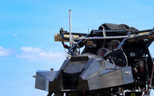 A heavily modified off-road utility vehicle (UTV/buggy) is stationary in an open grassy field under a clear blue sky. The vehicle has been extensively outfitted with what appears to be military or tactical equipment, including mounted systems on top, antennas, armor plating, and various electronic components. The vehicle appears to be an unmanned ground vehicle (UGV) or a remotely operated tactical platform.