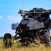 A heavily modified off-road utility vehicle (UTV/buggy) is stationary in an open grassy field under a clear blue sky. The vehicle has been extensively outfitted with what appears to be military or tactical equipment, including mounted systems on top, antennas, armor plating, and various electronic components. The vehicle appears to be an unmanned ground vehicle (UGV) or a remotely operated tactical platform.