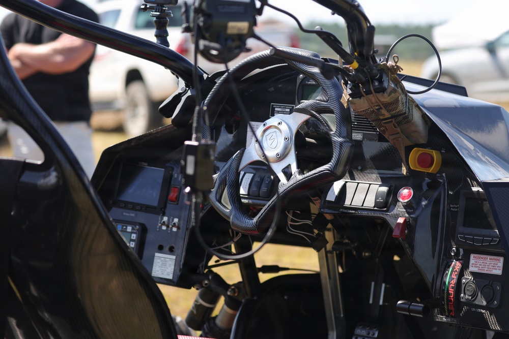 The image shows the interior cockpit of a high-performance or racing vehicle, featuring a carbon fiber steering wheel and a complex dashboard with multiple switches, buttons, and displays. The vehicle appears to be at an outdoor event or testing location, with people and other vehicles visible in the blurred background. The cockpit is heavily instrumented, suggesting a specialized racing, stunt, or experimental vehicle.