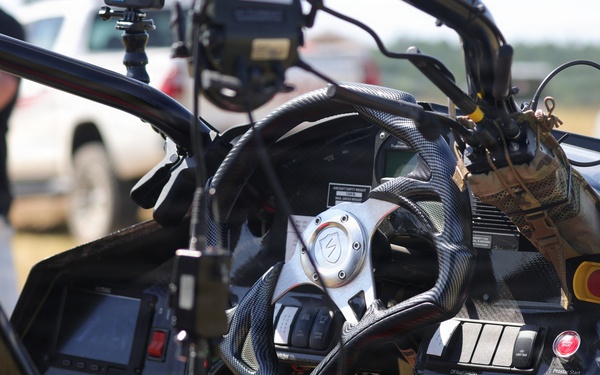The image shows the interior cockpit of a high-performance or racing vehicle, featuring a carbon fiber steering wheel and a complex dashboard with multiple switches, buttons, and displays. The vehicle appears to be at an outdoor event or testing location, with people and other vehicles visible in the blurred background. The cockpit is heavily instrumented, suggesting a specialized racing, stunt, or experimental vehicle.