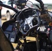 The image shows the interior cockpit of a high-performance or racing vehicle, featuring a carbon fiber steering wheel and a complex dashboard with multiple switches, buttons, and displays. The vehicle appears to be at an outdoor event or testing location, with people and other vehicles visible in the blurred background. The cockpit is heavily instrumented, suggesting a specialized racing, stunt, or experimental vehicle.