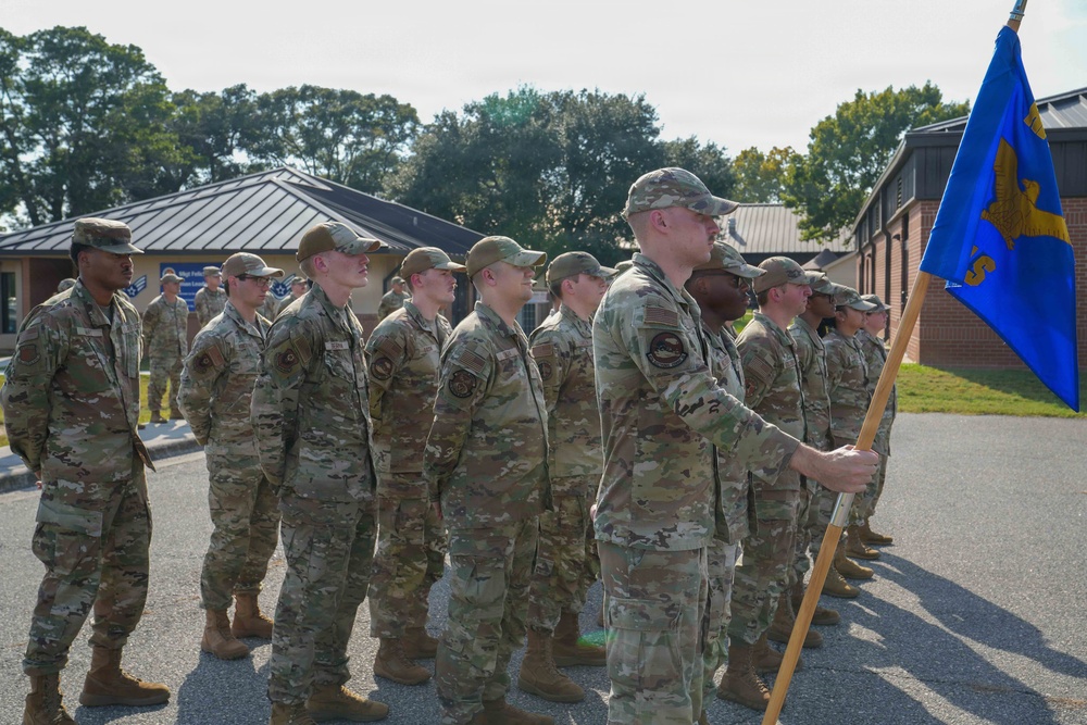 A formation of military personnel in OCP (Operational Camouflage Pattern) uniforms are standing at attention during what appears to be a formal military ceremony or parade. One service member in the foreground is holding a blue unit guidon flag with a gold emblem. The setting appears to be an outdoor paved area at a military installation with brick buildings and trees visible in the background.