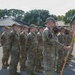 A formation of military personnel in OCP (Operational Camouflage Pattern) uniforms are standing at attention during what appears to be a formal military ceremony or parade. One service member in the foreground is holding a blue unit guidon flag with a gold emblem. The setting appears to be an outdoor paved area at a military installation with brick buildings and trees visible in the background.
