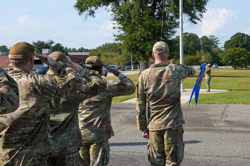 A group of military personnel in OCP (Operational Camouflage Pattern) uniforms are participating in a flag ceremony on a military installation. Several service members are rendering a salute while one individual holds a blue guidon or pennant near a flagpole. The setting appears to be an outdoor parade ground or common area on a military base during daytime.