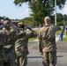 A group of military personnel in OCP (Operational Camouflage Pattern) uniforms are participating in a flag ceremony on a military installation. Several service members are rendering a salute while one individual holds a blue guidon or pennant near a flagpole. The setting appears to be an outdoor parade ground or common area on a military base during daytime.