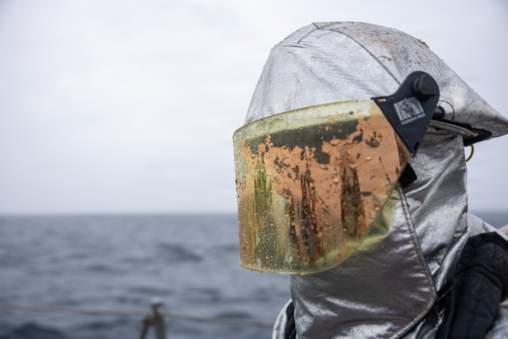 A person wearing a silver aluminized proximity/fire protection suit with a heavily soiled face shield is photographed against an overcast ocean backdrop. The visor shows significant contamination with orange and brown residue, likely from fire suppression foam, rust, or chemical exposure. The setting appears to be aboard a naval or maritime vessel.