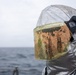 A person wearing a silver aluminized proximity/fire protection suit with a heavily soiled face shield is photographed against an overcast ocean backdrop. The visor shows significant contamination with orange and brown residue, likely from fire suppression foam, rust, or chemical exposure. The setting appears to be aboard a naval or maritime vessel.