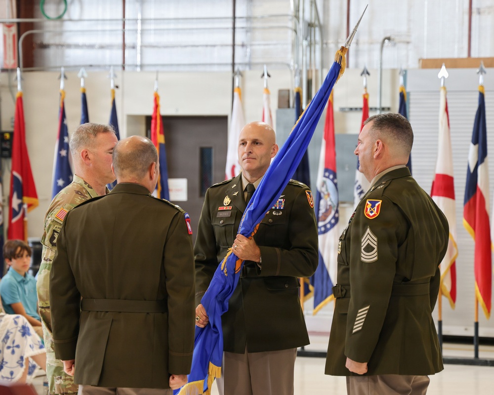 A U.S. Army change of command or activation ceremony is taking place inside what appears to be a military hangar or armory. Four military personnel in dress green and Army Combat Uniform (ACU) are participating in a guidon transfer, with one soldier holding a blue unit guidon/flag. Multiple state and military flags are displayed in the background.