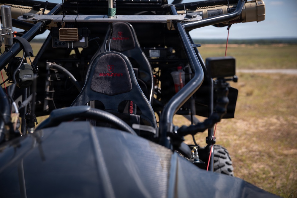 Close-up interior view of a tactical off-road vehicle, likely a military or special operations utility vehicle (UTV). The vehicle features carbon fiber bucket seats with branded headrests, a roll cage, and various mounted equipment including electronics and communication devices. The vehicle is positioned in an open field or training area visible in the background.