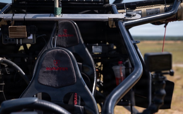 Close-up interior view of a tactical off-road vehicle, likely a military or special operations utility vehicle (UTV). The vehicle features carbon fiber bucket seats with branded headrests, a roll cage, and various mounted equipment including electronics and communication devices. The vehicle is positioned in an open field or training area visible in the background.