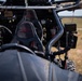 Close-up interior view of a tactical off-road vehicle, likely a military or special operations utility vehicle (UTV). The vehicle features carbon fiber bucket seats with branded headrests, a roll cage, and various mounted equipment including electronics and communication devices. The vehicle is positioned in an open field or training area visible in the background.