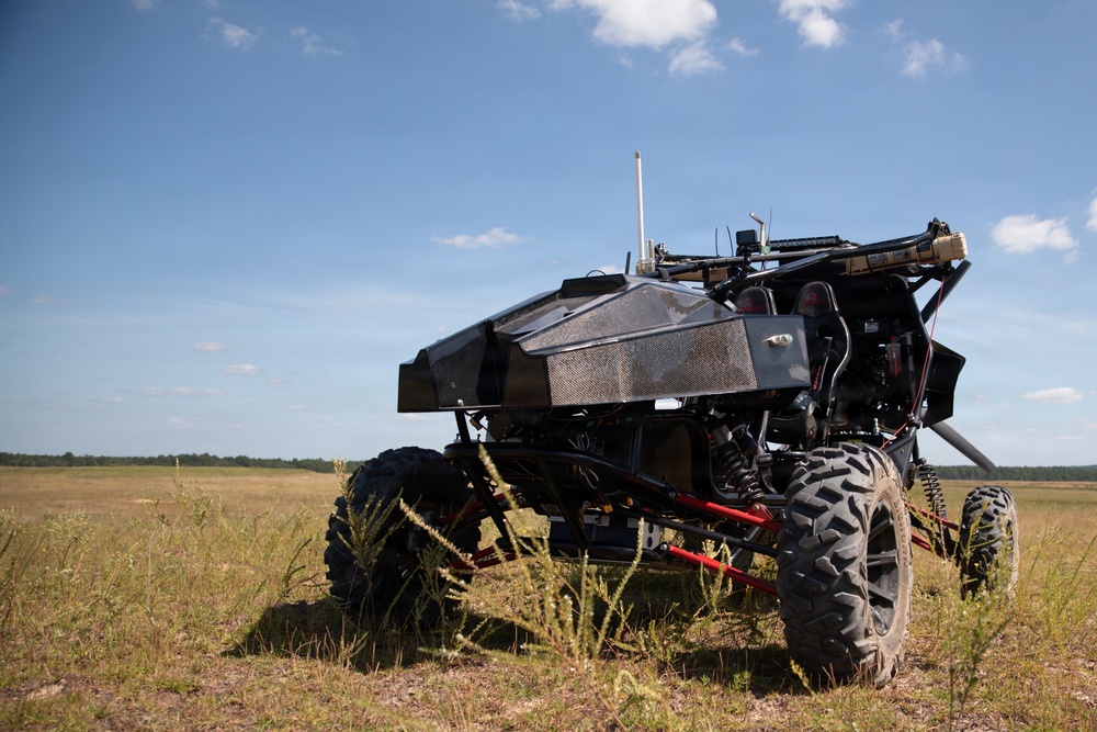 An unmanned ground vehicle (UGV) with a dark angular body shell is positioned in an open grassy field under a partly cloudy blue sky. The vehicle features large off-road tires, an exposed tubular frame with red components, and multiple sensors or antennas mounted on top. The setting appears to be a military or testing range environment.