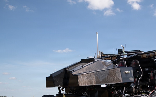 An unmanned ground vehicle (UGV) with a dark angular body shell is positioned in an open grassy field under a partly cloudy blue sky. The vehicle features large off-road tires, an exposed tubular frame with red components, and multiple sensors or antennas mounted on top. The setting appears to be a military or testing range environment.