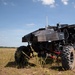 An unmanned ground vehicle (UGV) with a dark angular body shell is positioned in an open grassy field under a partly cloudy blue sky. The vehicle features large off-road tires, an exposed tubular frame with red components, and multiple sensors or antennas mounted on top. The setting appears to be a military or testing range environment.