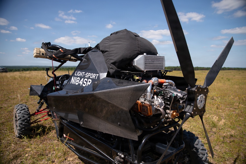 The image shows a light-sport aircraft or powered parachute/paramotor buggy with a carbon fiber body, registration number N184SR, parked on an open grassy field. The vehicle features a rear-mounted pusher propeller, a visible engine with turbocharger components, and an intercooler, with what appears to be a parachute or canopy stored in a black bag on top. The setting is a rural airstrip or open field under partly cloudy skies.
