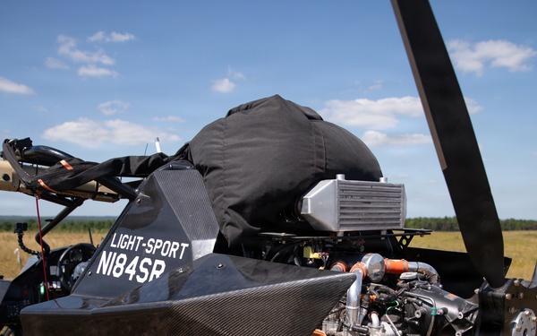 The image shows a light-sport aircraft or powered parachute/paramotor buggy with a carbon fiber body, registration number N184SR, parked on an open grassy field. The vehicle features a rear-mounted pusher propeller, a visible engine with turbocharger components, and an intercooler, with what appears to be a parachute or canopy stored in a black bag on top. The setting is a rural airstrip or open field under partly cloudy skies.