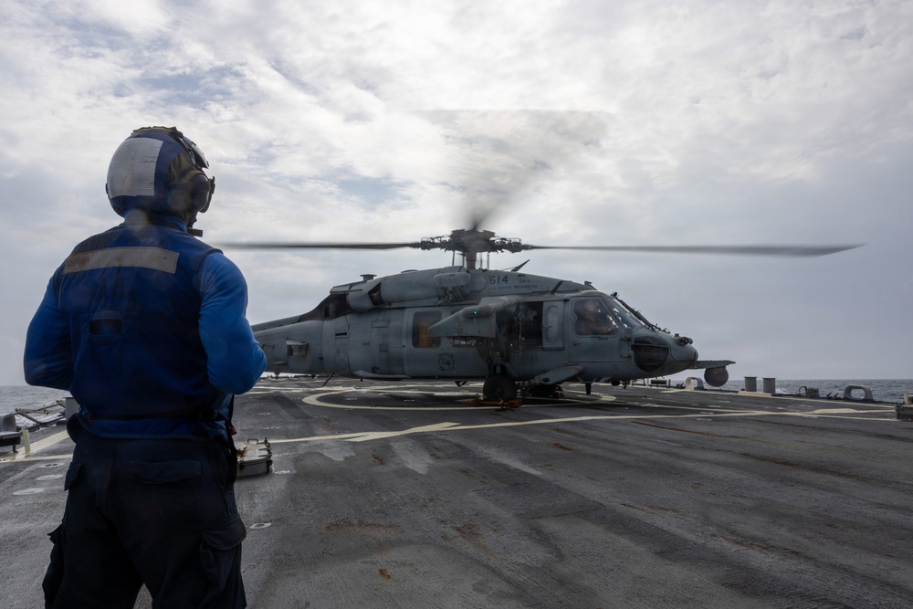A naval flight deck crew member in a blue uniform and helmet stands on a ship's flight deck observing a military helicopter with spinning rotors. The helicopter, marked with the number '614', is positioned on the landing pad of what appears to be a U.S. Navy vessel at sea. The scene depicts a standard flight deck operation with the helicopter either preparing for takeoff or having just landed.