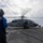 A naval flight deck crew member in a blue uniform and helmet stands on a ship's flight deck observing a military helicopter with spinning rotors. The helicopter, marked with the number '614', is positioned on the landing pad of what appears to be a U.S. Navy vessel at sea. The scene depicts a standard flight deck operation with the helicopter either preparing for takeoff or having just landed.