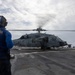A naval flight deck crew member in a blue uniform and helmet stands on a ship's flight deck observing a military helicopter with spinning rotors. The helicopter, marked with the number '614', is positioned on the landing pad of what appears to be a U.S. Navy vessel at sea. The scene depicts a standard flight deck operation with the helicopter either preparing for takeoff or having just landed.