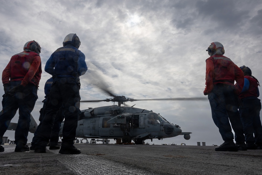 Naval flight deck personnel observe a military helicopter with spinning rotors on what appears to be a ship's flight deck at sea. The crew members are wearing flight deck safety gear including helmets and colored jerseys indicating their roles. The scene is set under an overcast sky with the helicopter appearing ready for launch or having just landed.