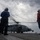 Naval flight deck personnel observe a military helicopter with spinning rotors on what appears to be a ship's flight deck at sea. The crew members are wearing flight deck safety gear including helmets and colored jerseys indicating their roles. The scene is set under an overcast sky with the helicopter appearing ready for launch or having just landed.