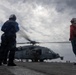 Naval flight deck personnel observe a military helicopter with spinning rotors on what appears to be a ship's flight deck at sea. The crew members are wearing flight deck safety gear including helmets and colored jerseys indicating their roles. The scene is set under an overcast sky with the helicopter appearing ready for launch or having just landed.