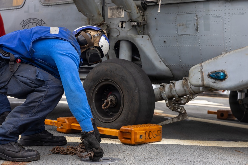 A naval deck crew member in blue coveralls and a white helmet is chocking and chaining a helicopter on what appears to be a ship's flight deck. The orange wheel chocks are marked 'DDG 53', identifying this as the USS John Paul Jones, an Arleigh Burke-class destroyer. The worker is inspecting or securing the landing gear area of a gray military helicopter.