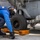 A naval deck crew member in blue coveralls and a white helmet is chocking and chaining a helicopter on what appears to be a ship's flight deck. The orange wheel chocks are marked 'DDG 53', identifying this as the USS John Paul Jones, an Arleigh Burke-class destroyer. The worker is inspecting or securing the landing gear area of a gray military helicopter.