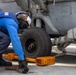A naval deck crew member in blue coveralls and a white helmet is chocking and chaining a helicopter on what appears to be a ship's flight deck. The orange wheel chocks are marked 'DDG 53', identifying this as the USS John Paul Jones, an Arleigh Burke-class destroyer. The worker is inspecting or securing the landing gear area of a gray military helicopter.