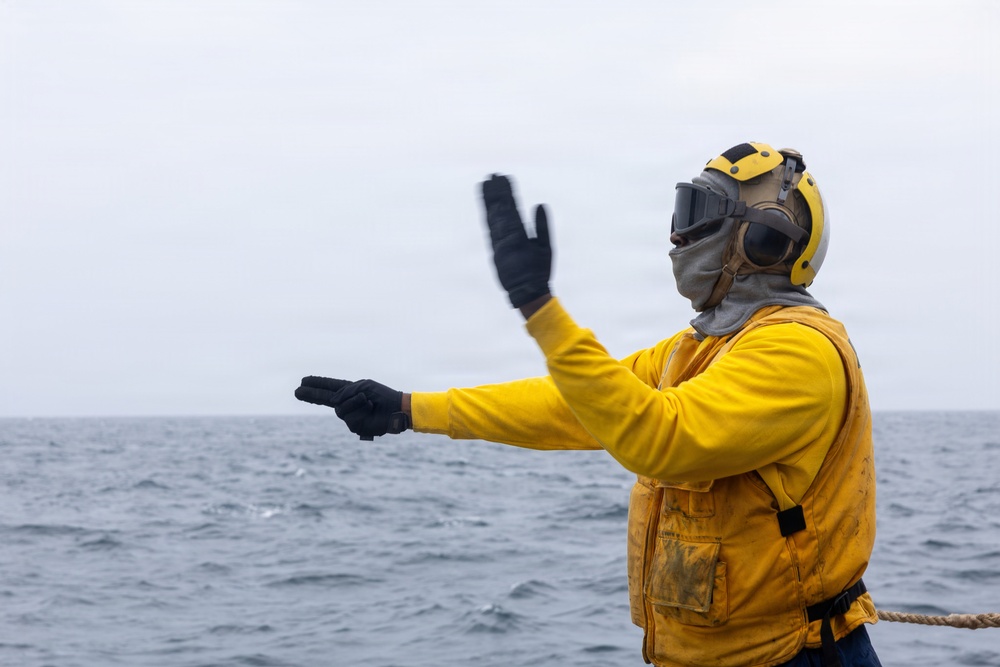 A person wearing yellow flight deck gear, including a helmet with ear protection, goggles, a balaclava, and black gloves, is giving hand signals over open ocean. The individual appears to be a naval flight deck crew member, identifiable by the yellow jersey and protective equipment. The overcast sky and choppy gray water suggest maritime operational conditions.