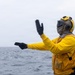 A person wearing yellow flight deck gear, including a helmet with ear protection, goggles, a balaclava, and black gloves, is giving hand signals over open ocean. The individual appears to be a naval flight deck crew member, identifiable by the yellow jersey and protective equipment. The overcast sky and choppy gray water suggest maritime operational conditions.