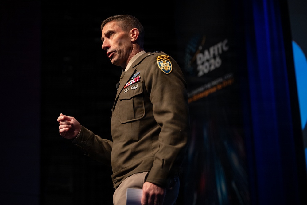 A U.S. Army officer in dress uniform is speaking at a conference or event identified as 'DAFITC 2026' visible on the backdrop. The officer is gesturing with a raised fist while delivering a presentation on stage. Multiple military insignia, patches, and medals are visible on the uniform.