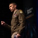 A U.S. Army officer in dress uniform is speaking at a conference or event identified as 'DAFITC 2026' visible on the backdrop. The officer is gesturing with a raised fist while delivering a presentation on stage. Multiple military insignia, patches, and medals are visible on the uniform.