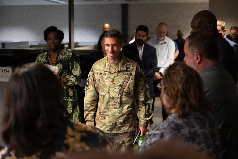 A U.S. Army soldier identified by name tape as 'STANTON' is engaged in conversation with a group of civilians in what appears to be an indoor meeting or conference setting. The soldier is wearing OCP (Operational Camouflage Pattern) uniform with 'U.S. ARMY' tape visible and rank insignia on the chest. The gathering appears to be a professional or community engagement event with a diverse group of attendees.