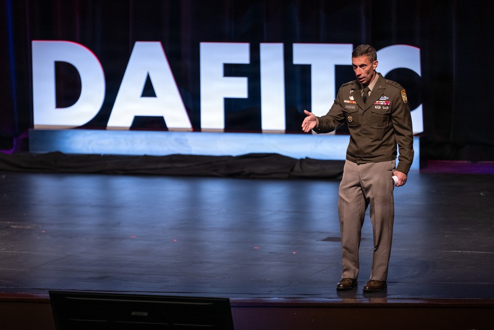 A military officer in olive green dress uniform is speaking on a stage at what appears to be a DAFITC (Defense Acquisition Functional Integrated Training Course) event. The officer is gesturing with an outstretched hand, suggesting an active presentation or keynote address. The backdrop features large illuminated white letters spelling 'DAFITC' against a dark stage setting.