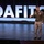 Lt. Gen. Stanton Speaks at DAFTIC A military officer in olive green dress uniform is speaking on a stage at what appears to be a DAFITC (Defense Acquisition Functional Integrated Training Course) event. The officer is gesturing with an outstretched hand, suggesting an active presentation or keynote address. The backdrop features large illuminated white letters spelling 'DAFITC' against a dark stage setting.