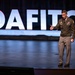 A military officer in olive green dress uniform is speaking on a stage at what appears to be a DAFITC (Defense Acquisition Functional Integrated Training Course) event. The officer is gesturing with an outstretched hand, suggesting an active presentation or keynote address. The backdrop features large illuminated white letters spelling 'DAFITC' against a dark stage setting.