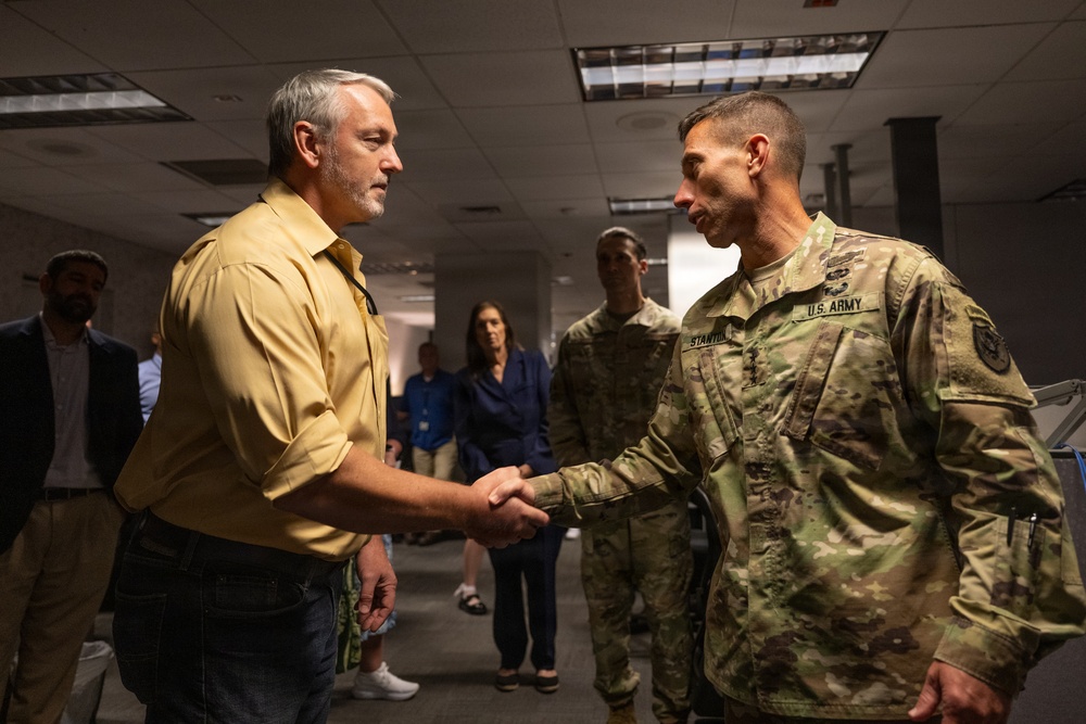 A civilian man in a yellow shirt is shaking hands with a U.S. Army soldier in OCP (Operational Camouflage Pattern) uniform. The meeting appears to be taking place in an office or government building environment. Several other individuals are visible in the background observing the exchange.