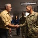 A civilian man in a yellow shirt is shaking hands with a U.S. Army soldier in OCP (Operational Camouflage Pattern) uniform. The meeting appears to be taking place in an office or government building environment. Several other individuals are visible in the background observing the exchange.