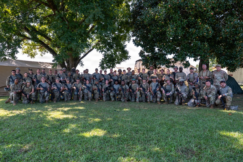 A large group of military personnel in camouflage uniforms pose for a formal unit photograph outdoors beneath large trees. The group consists of approximately 50-60 soldiers arranged in two to three rows, many holding rifles or wearing tactical gear. The setting appears to be a military installation or training area with tents and structures visible in the background.